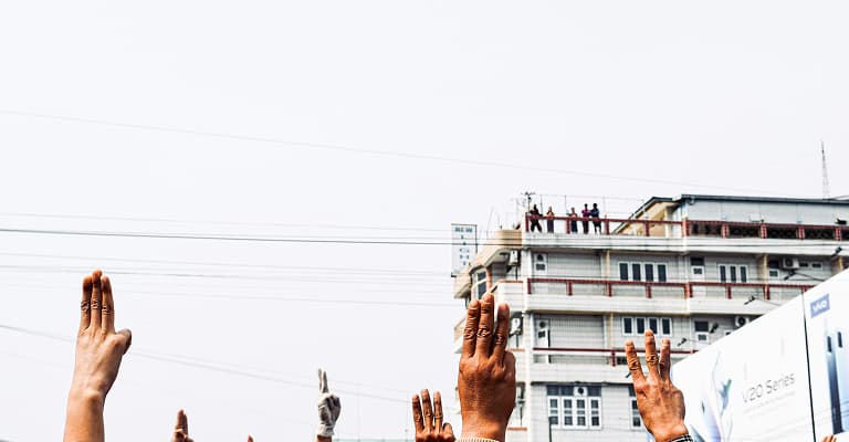 Hands raised in protest with three-finger salute, against a backdrop of a building, symbolizing unity and resistance.